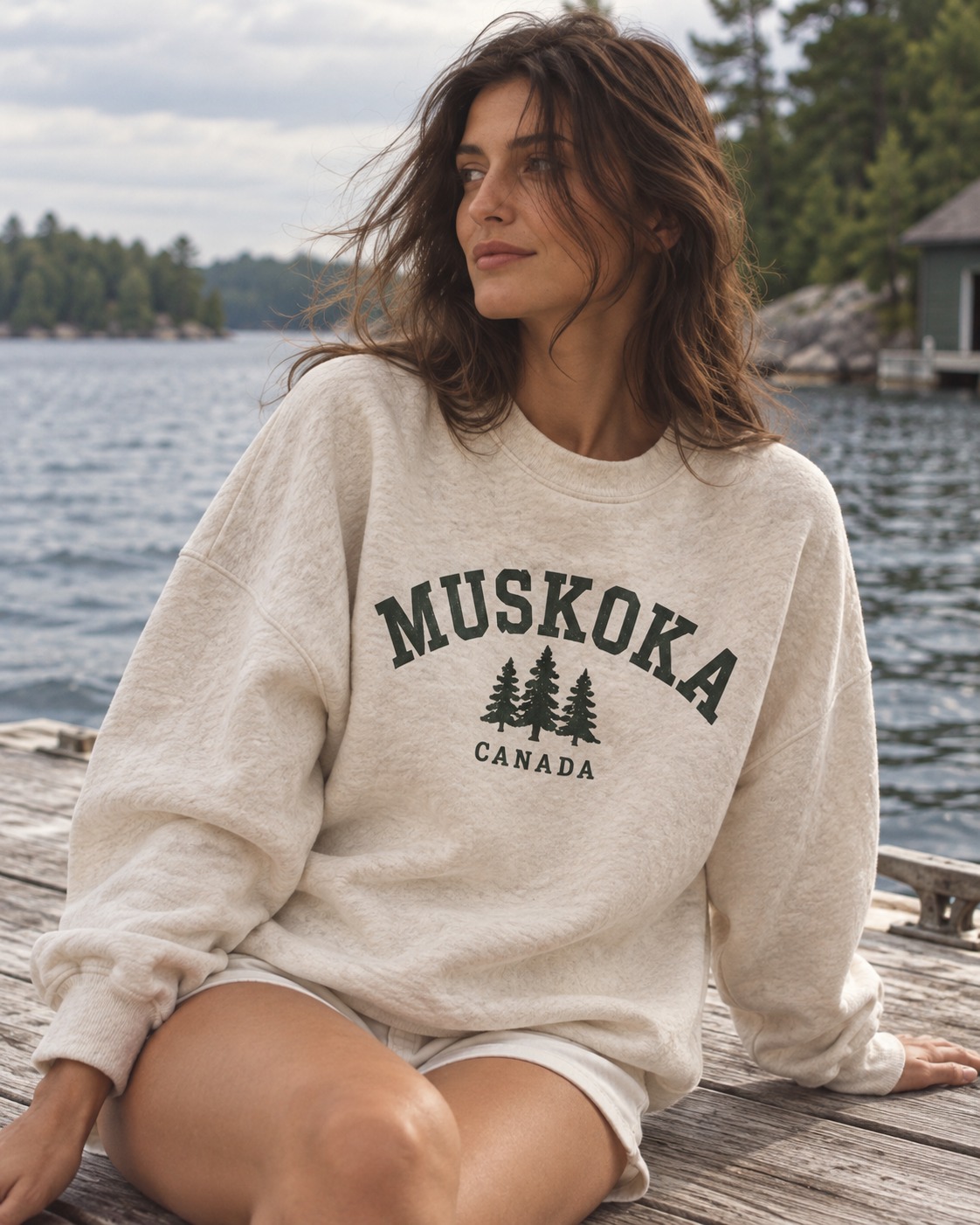 Oatmeal Muskoka Canada crewneck with forest-green lettering and pines, worn on a lakeside dock